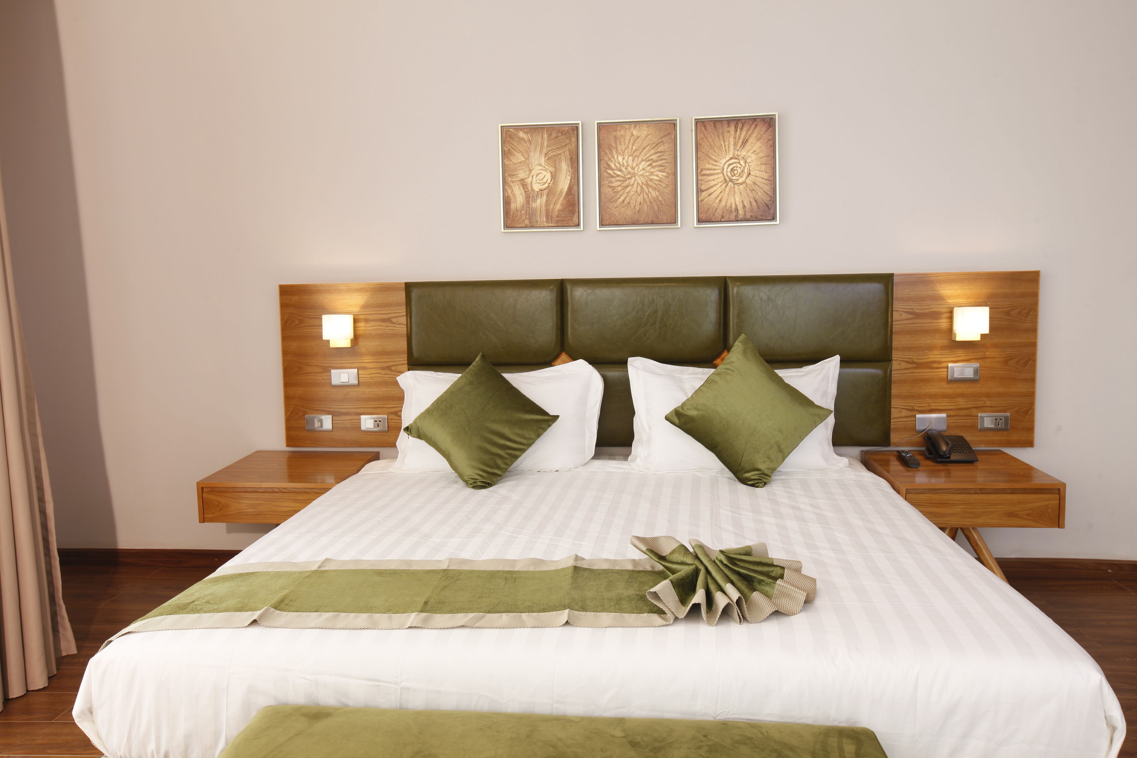 Een netjes opgemaakt bed met een groen sierkussen, een nachtkastje met een telefoon, en een wandlamp.