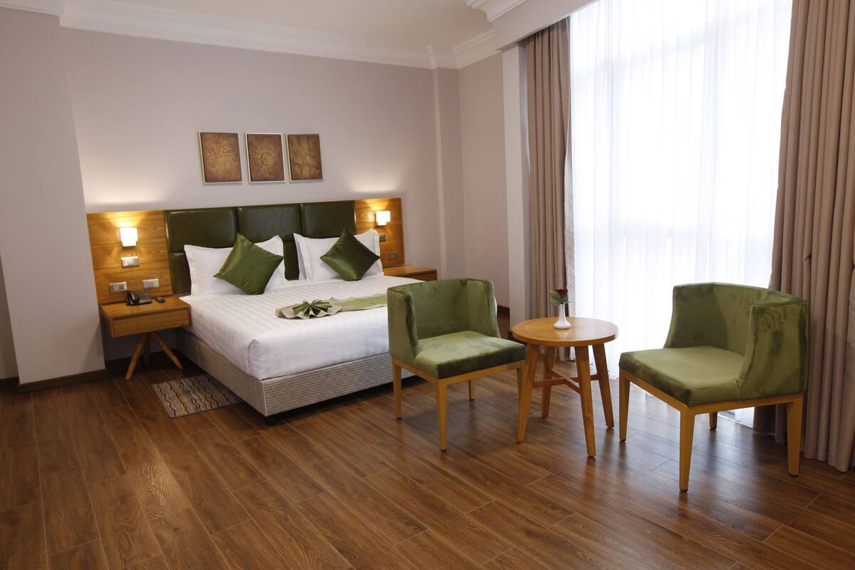 A hotel room with a bed, two green armchairs, a small round table, wooden flooring, and a nightstand with a lamp.
