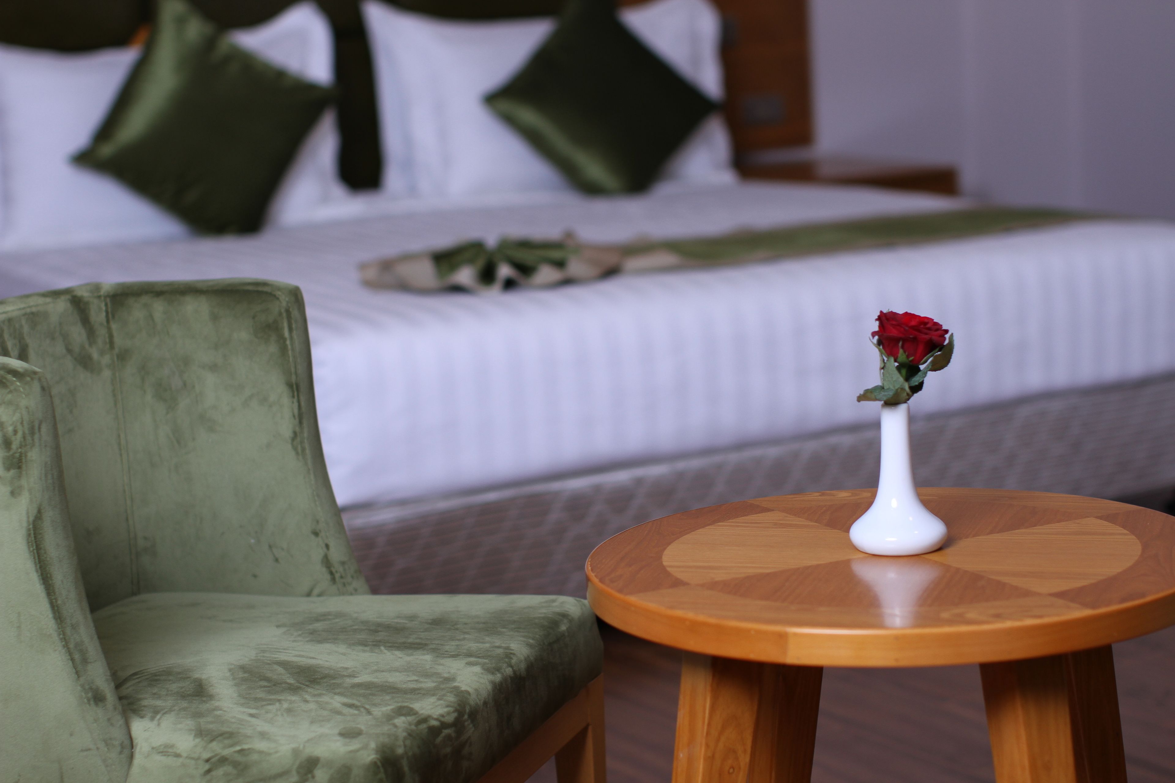 A hotel room with a bed, a green armchair, and a small wooden table with a vase of a red rose.