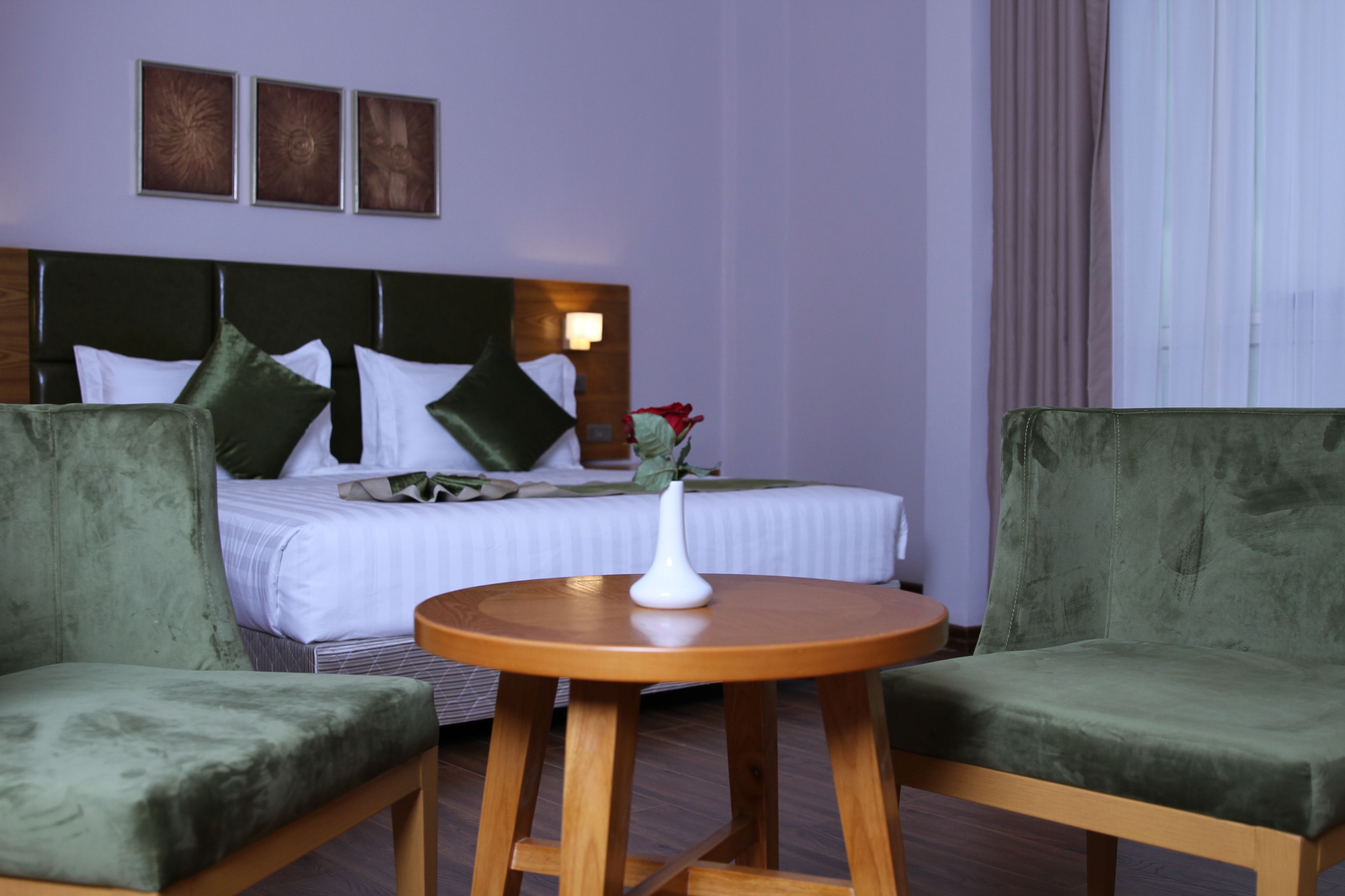 Una habitación de hotel con una cama, dos sillones verdes, una mesa de madera y un florero con una flor.