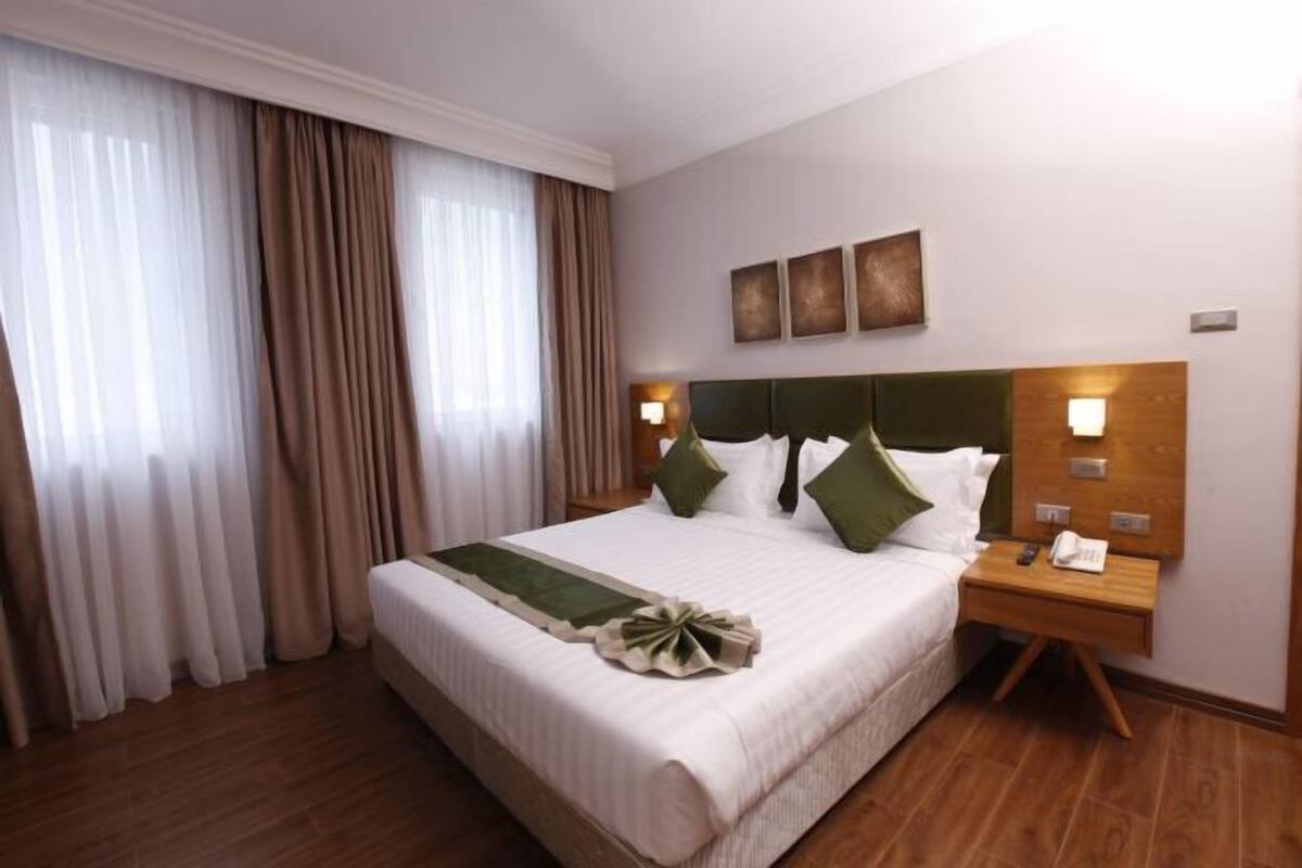 A hotel room with a large bed, a wooden nightstand, a wall-mounted phone, and a window with curtains.