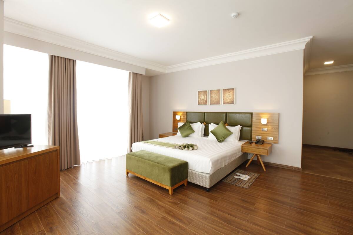 A modern hotel room with a large bed, a wooden desk, a television, and a large window with curtains.