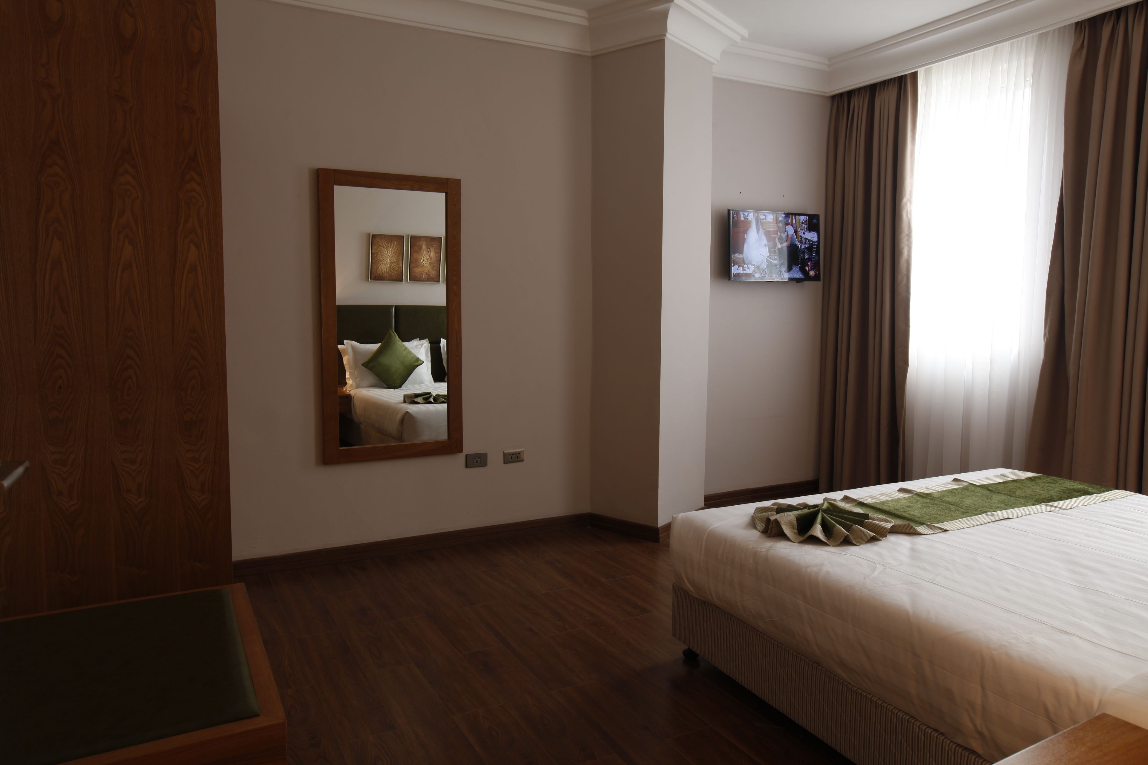 Une chambre d’hôtel avec un grand lit, une armoire en bois, un miroir, une télévision et une fenêtre avec des rideaux.