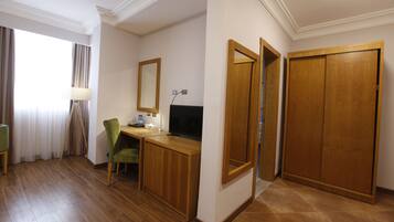 A hotel room with a wooden desk, a green chair, a television, a mirror, and a wardrobe.