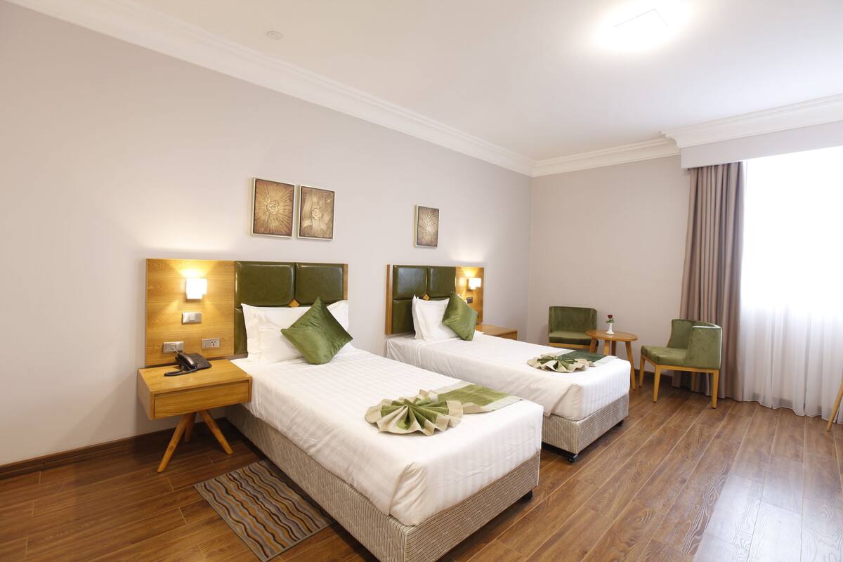 A hotel room with two beds, a small table, a chair, and a window with curtains.