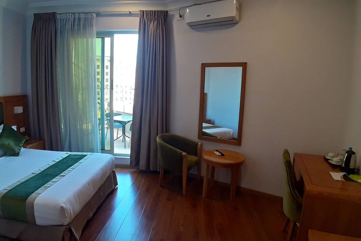 A hotel room with a bed, a desk, a chair, a mirror, and a view of the outside.