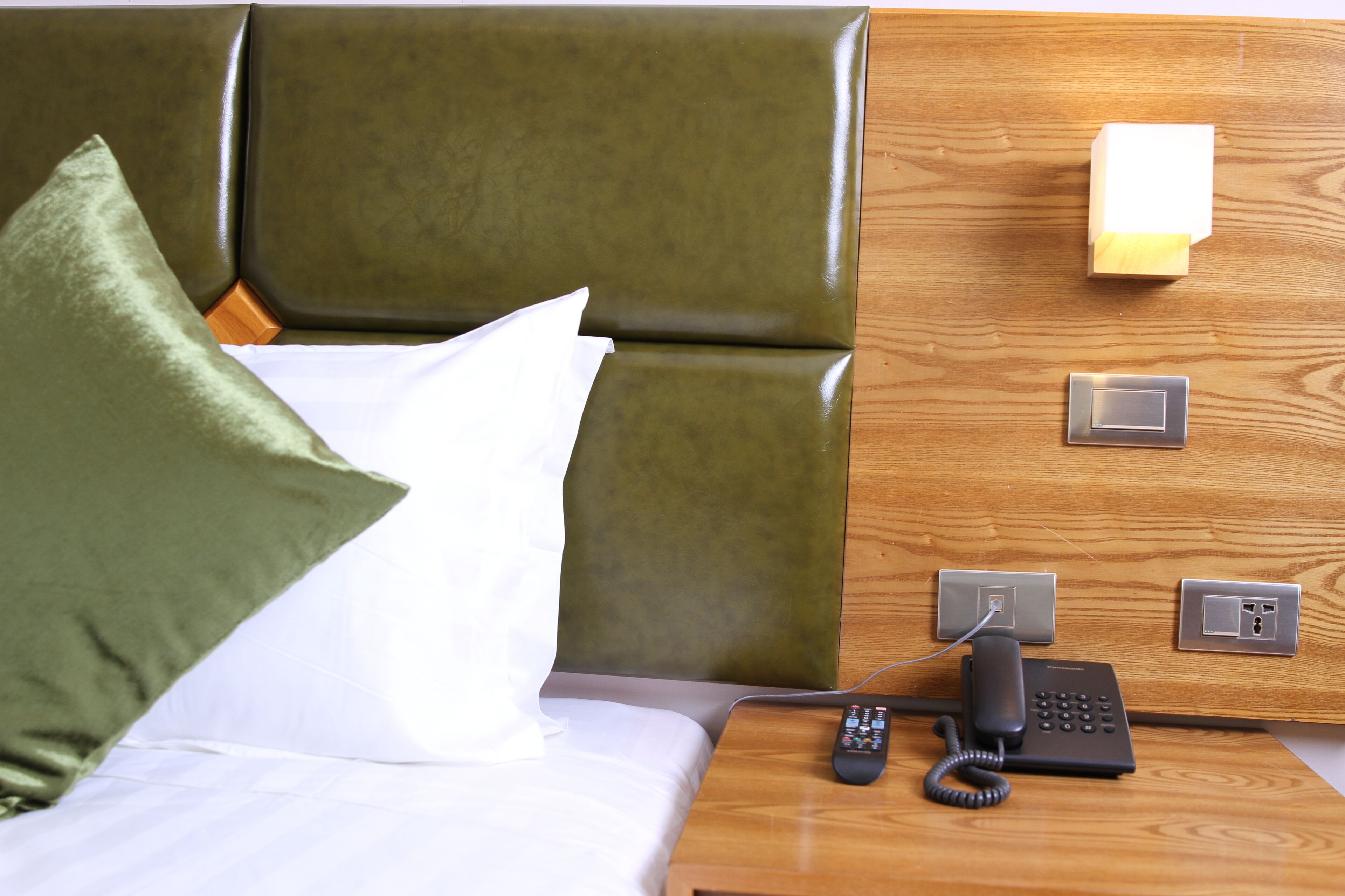 A hotel room with a green upholstered headboard, a white pillow, a green pillow, a wooden nightstand with a telephone and power outlets.