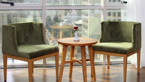 Een kamer met twee groene fauteuils, een houten tafel met een vaas en een roos, en grote ramen met uitzicht op de stad.