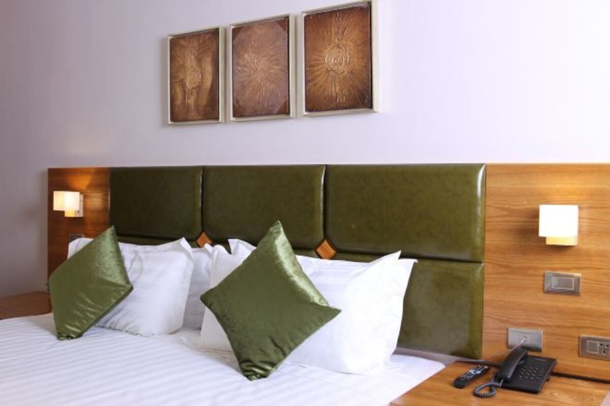 Una habitación de hotel con cama, cabecera verde, ropa de cama blanca y un teléfono en la mesita de noche.