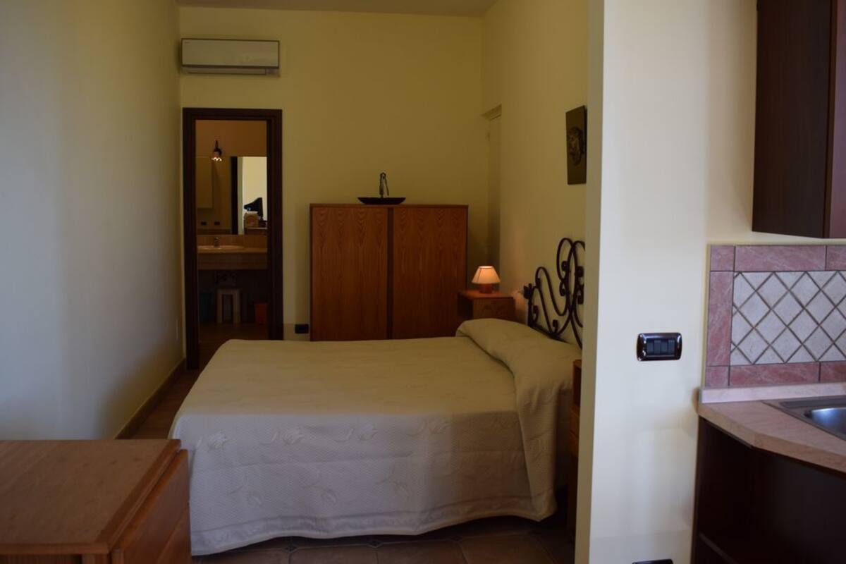 A bedroom with a bed, a wooden wardrobe, a bedside table with a lamp, and a small desk.