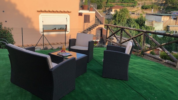 A terrace with wicker furniture, a small table, and a view of a hillside with buildings. - Milord Relais (Massa Lubrense)