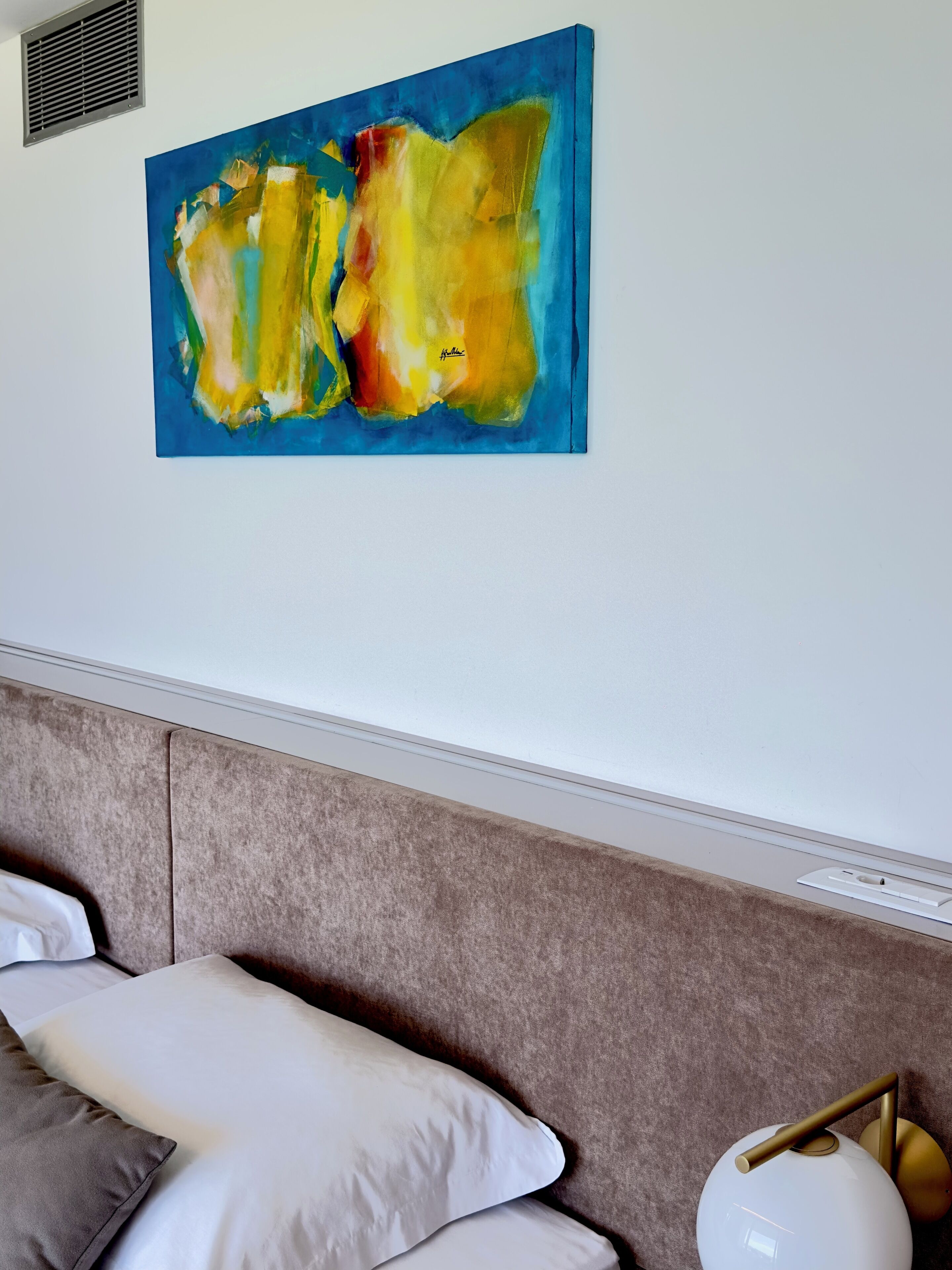 A room with a large abstract painting, a bed with a headboard, and a bedside lamp.