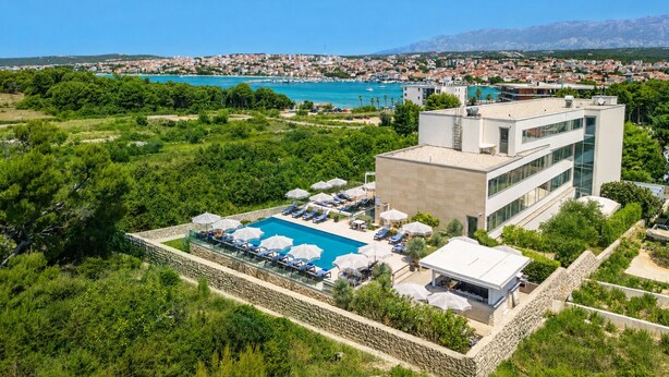 Un hotel moderno con piscina, rodeado de vegetación y vistas a un pueblo costero.