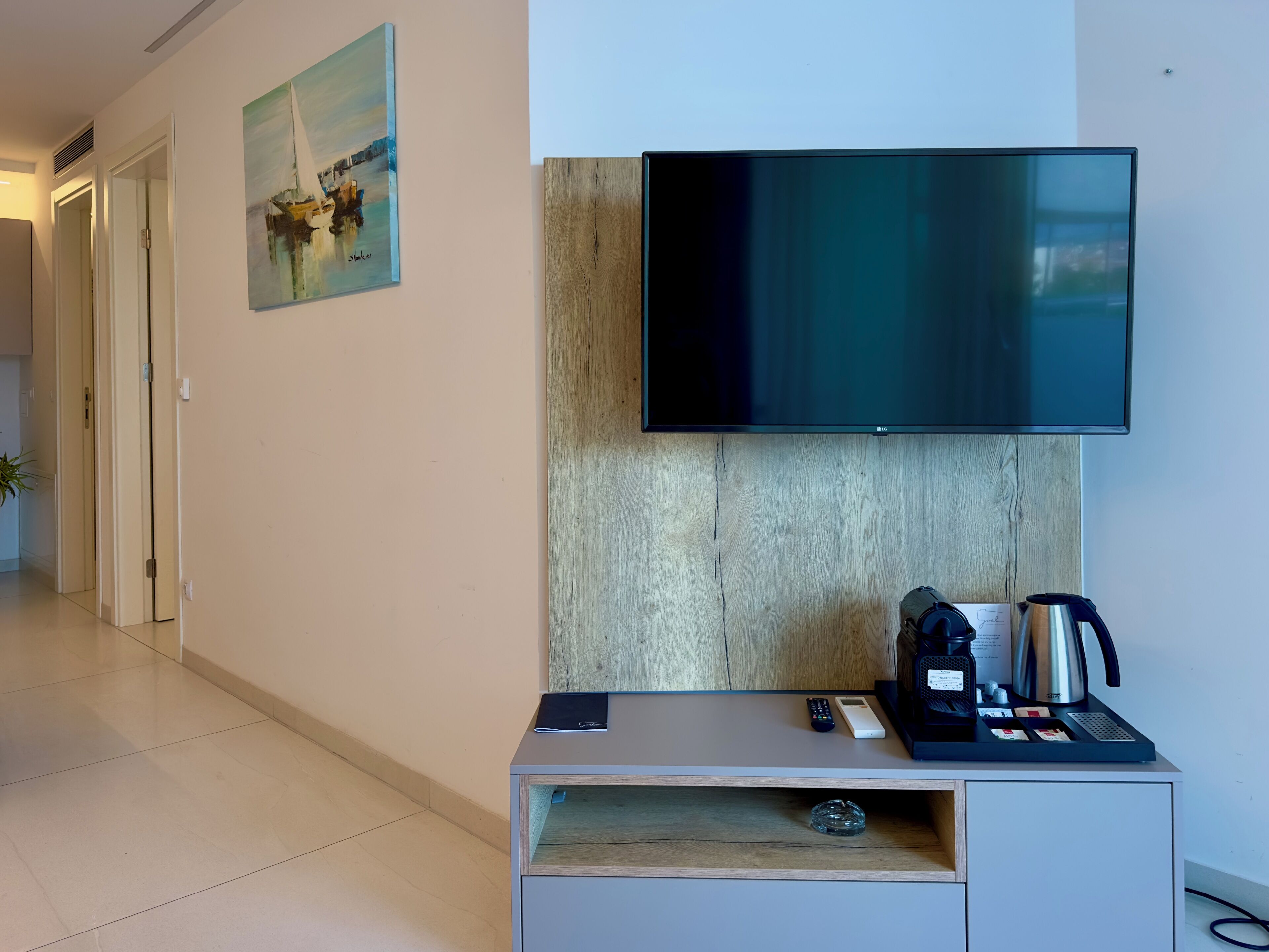 A modern hotel room with a flat-screen TV, a coffee maker, a remote control, and a small shelf with a glass container.