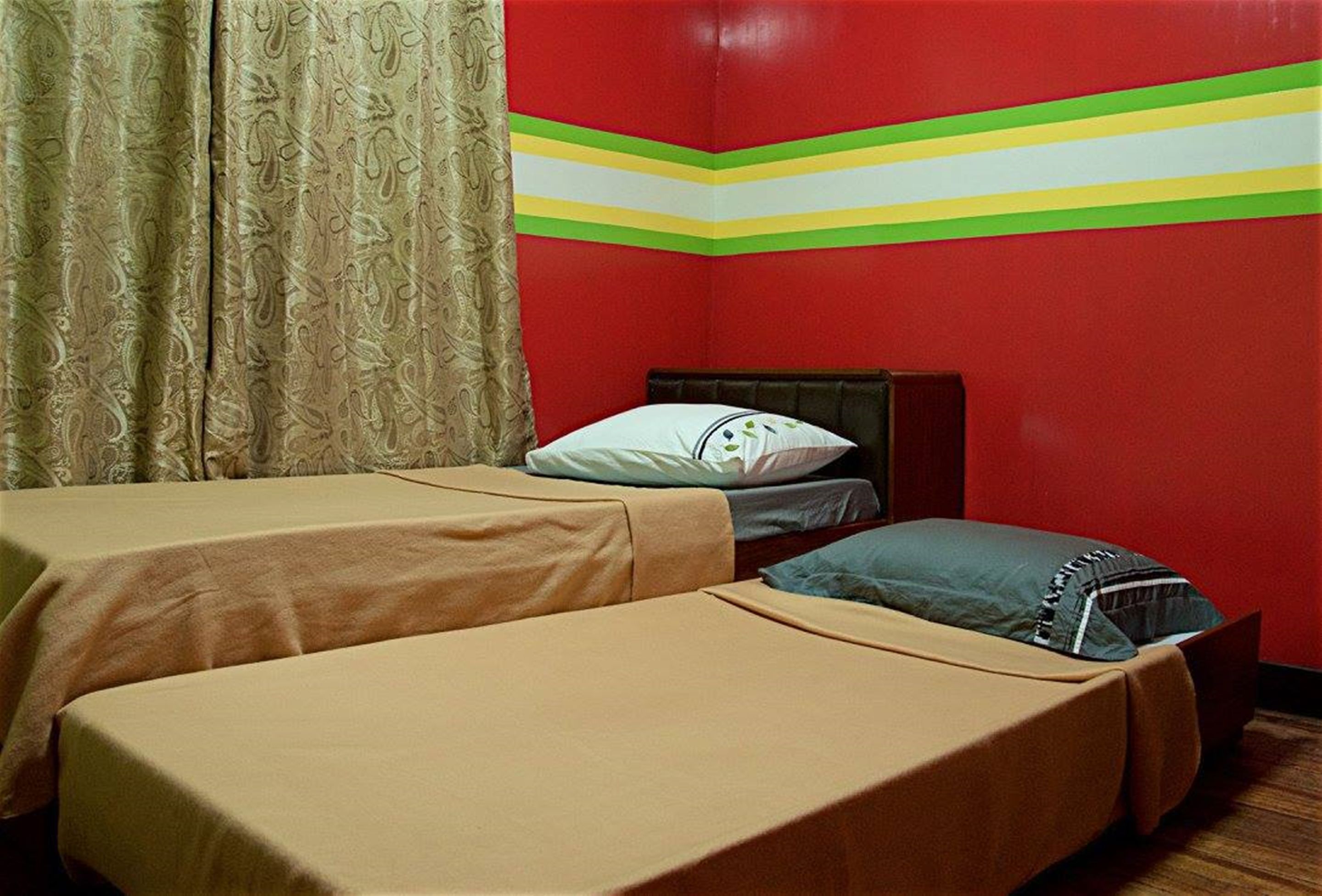 A room with two single beds, a wooden headboard, a patterned curtain, and a red wall with a green and yellow stripe.