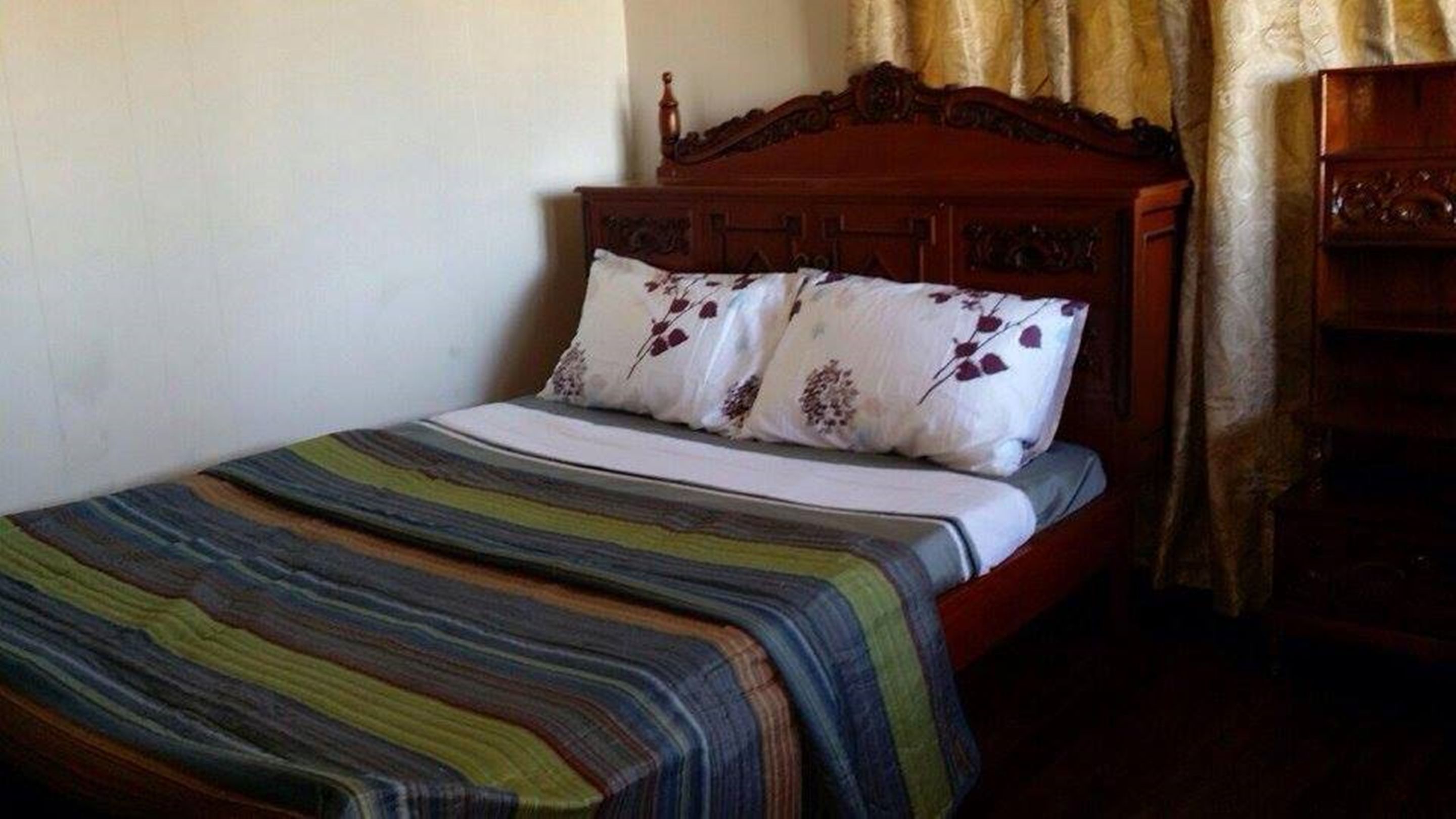 A single bed with a striped bedspread and two pillows with floral patterns, a wooden headboard, and a wooden nightstand with a lamp.