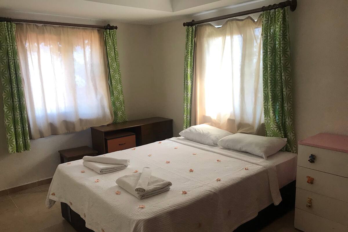 A neatly made bed with white linens and a floral-patterned bedspread, a bedside table, a nightstand, and a dresser with drawers.