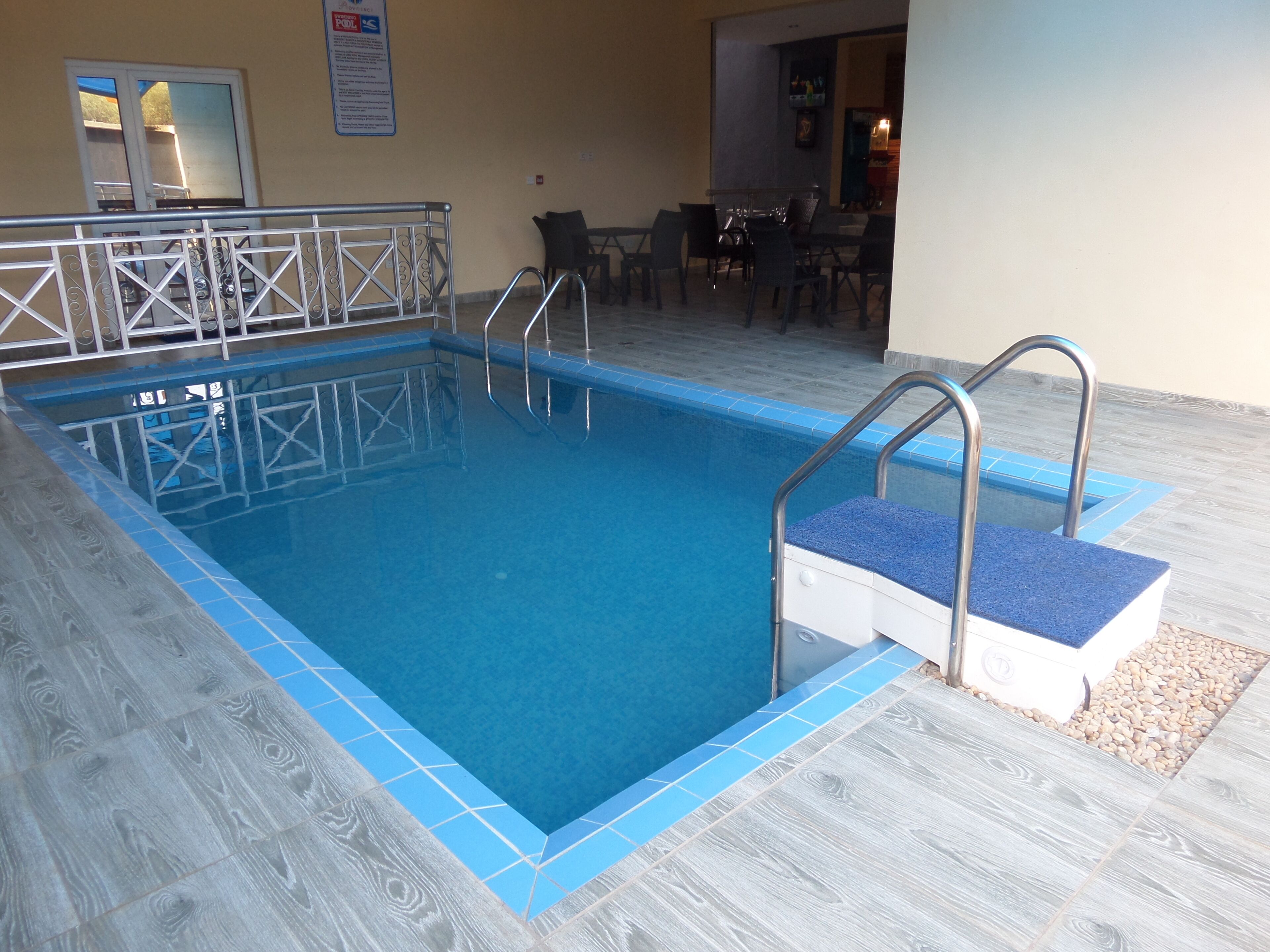 An indoor swimming pool with a blue tiled edge, a metal ladder, and a seating area with chairs and tables.