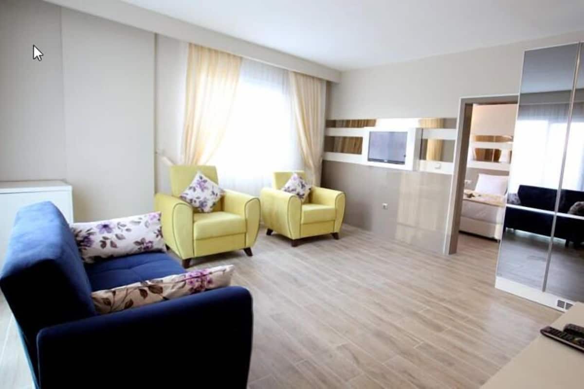 A modern hotel room with a blue sofa, two yellow armchairs, a large mirror, and a bed in the background.