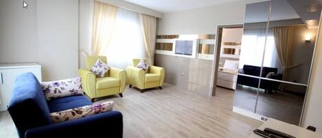 Habitación de hotel moderna con un sofá azul, dos sillones amarillos, un espejo grande y una cama al fondo.
