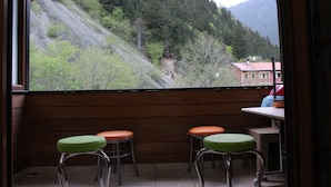 Een kamer met een groot raam met uitzicht op een bergachtig landschap. Er staan twee krukken met groene kussens bij een tafel waarop een rood object ligt.