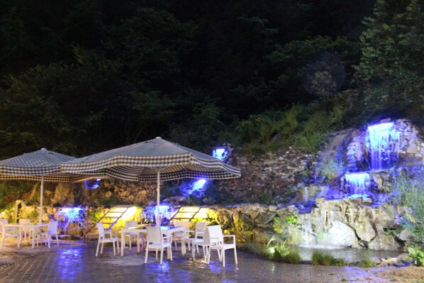 An outdoor dining area with tables and chairs under checkered umbrellas, a waterfall, and a stone wall backdrop. - Selale Otel ve Bungalow (Çaykara)