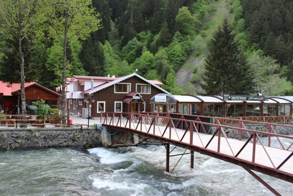 Una cabaña de madera con techo rojo, un puente sobre un río y un paisaje montañoso boscoso al fondo.