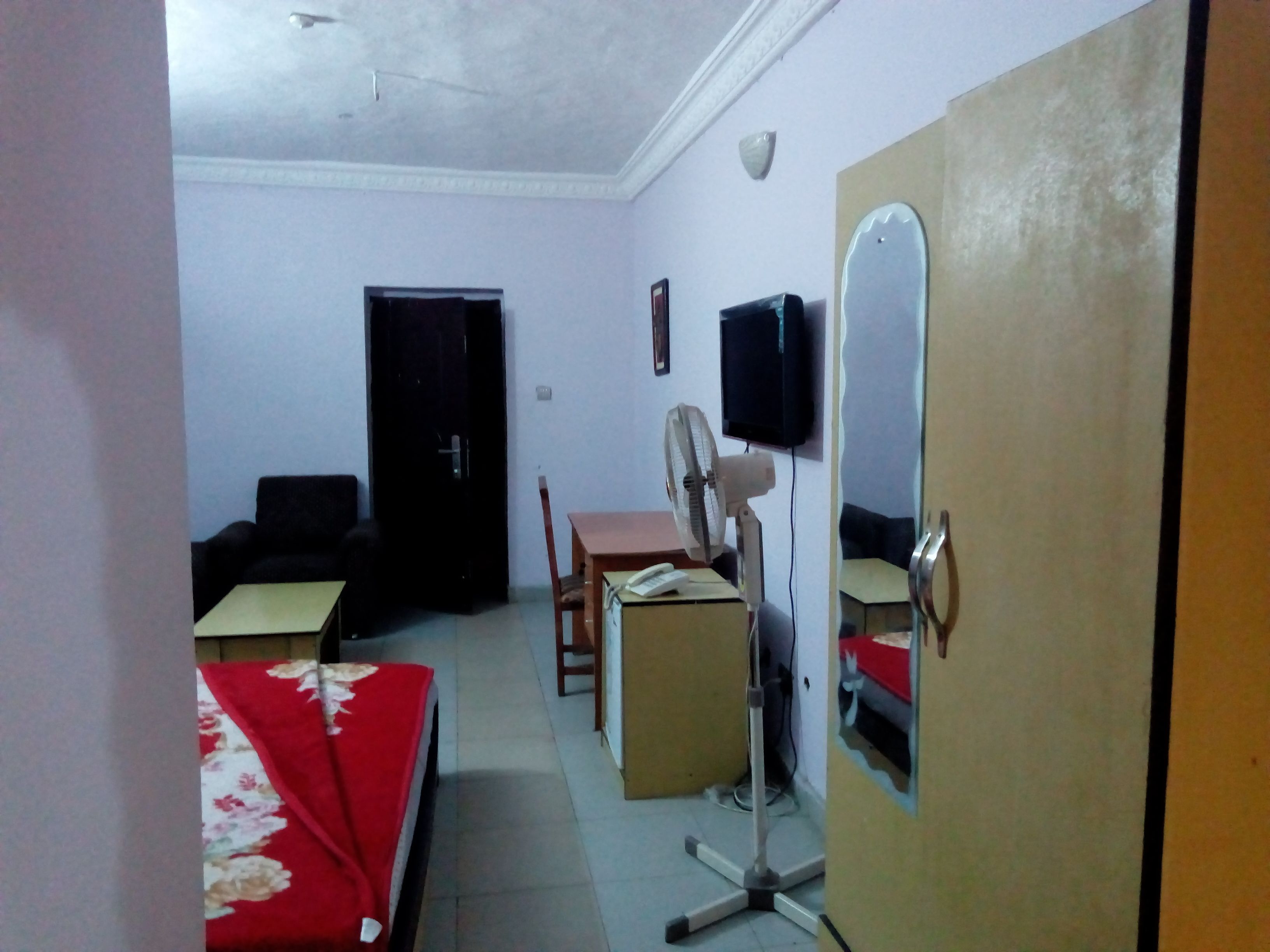 A room with a bed, a desk, a chair, a fan, a television, and a door.