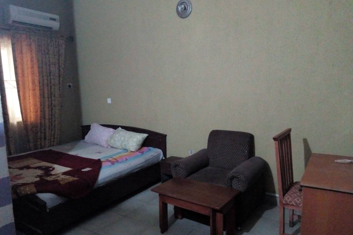 A bedroom with a bed, a chair, a small table, and an air conditioning unit on the wall.