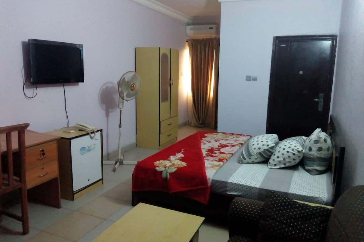 A compact hotel room with a bed, a desk, a chair, a wardrobe, a television, and a standing fan.