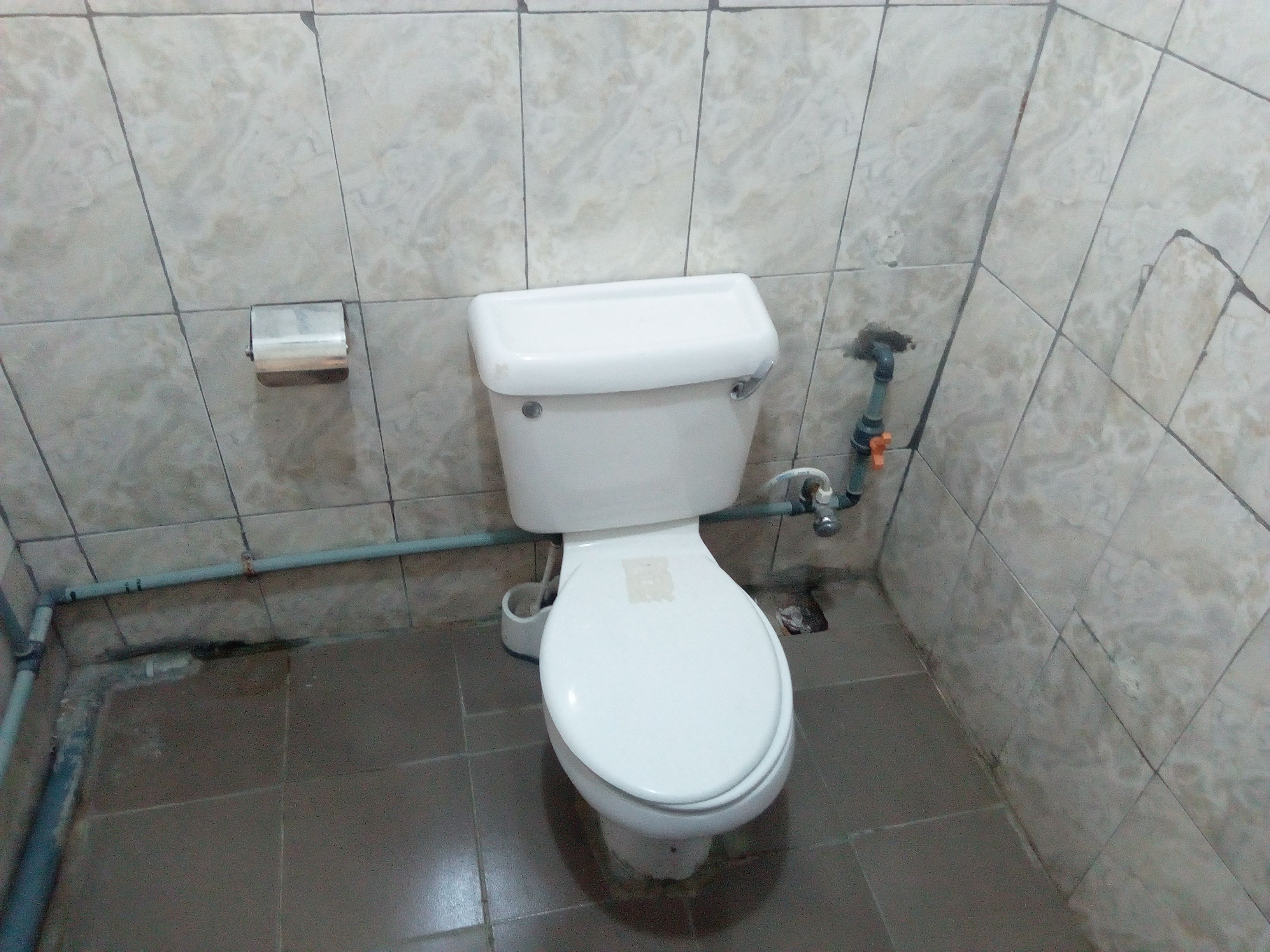 A standard white toilet with a tank and a seat, a visible pipe, and a small shelf on the wall.