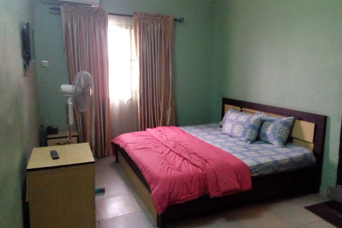 A bedroom with a bed, a standing fan, a desk, and a window with curtains.