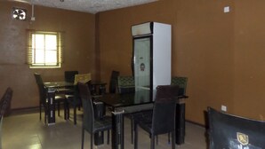 A dining area with a glass table and chairs, a white refrigerator, and a window with blinds. - Prowess Hotel and Suites (Ota)