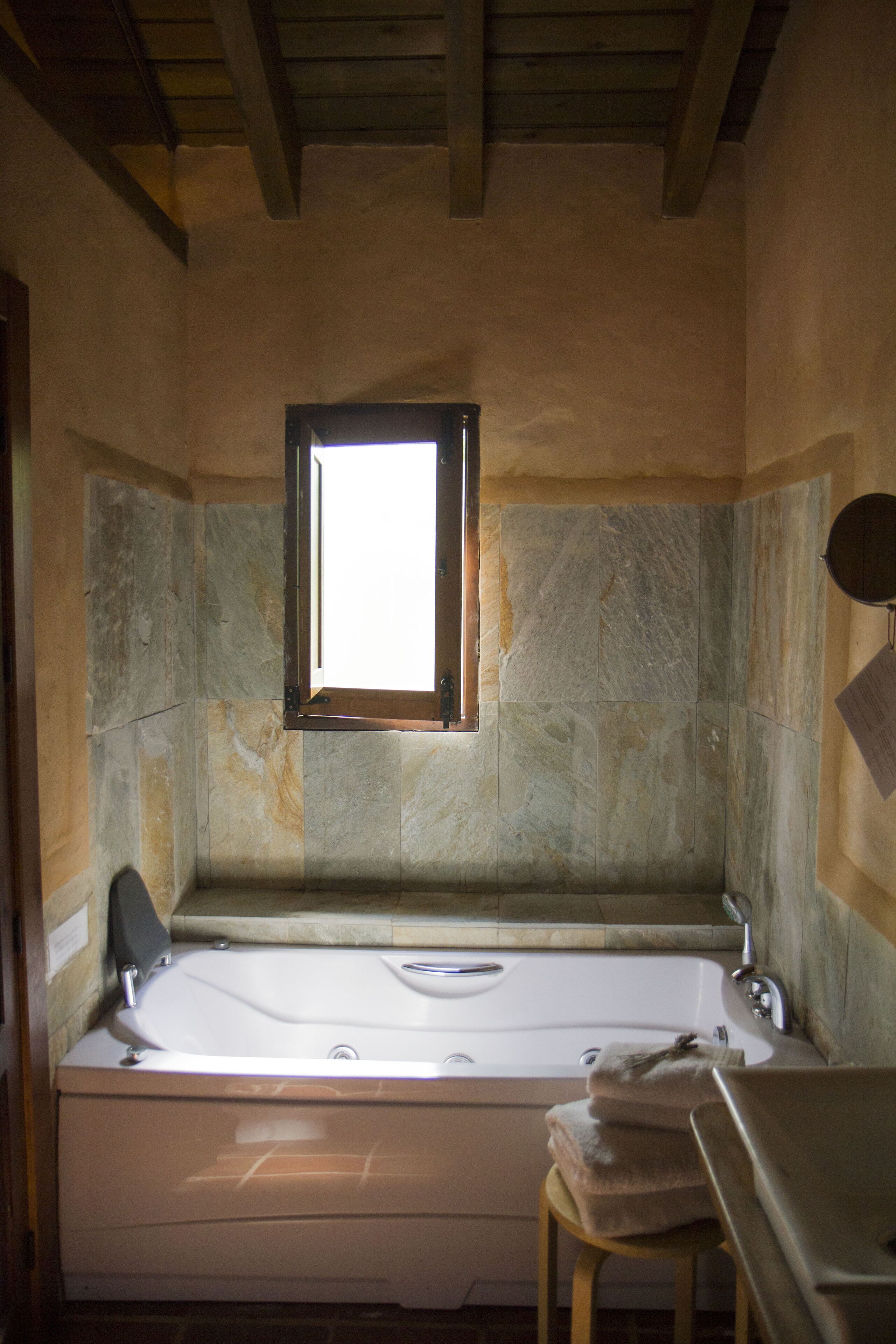 Un baño con pared de piedra, una ventana, una bañera y un taburete de madera.