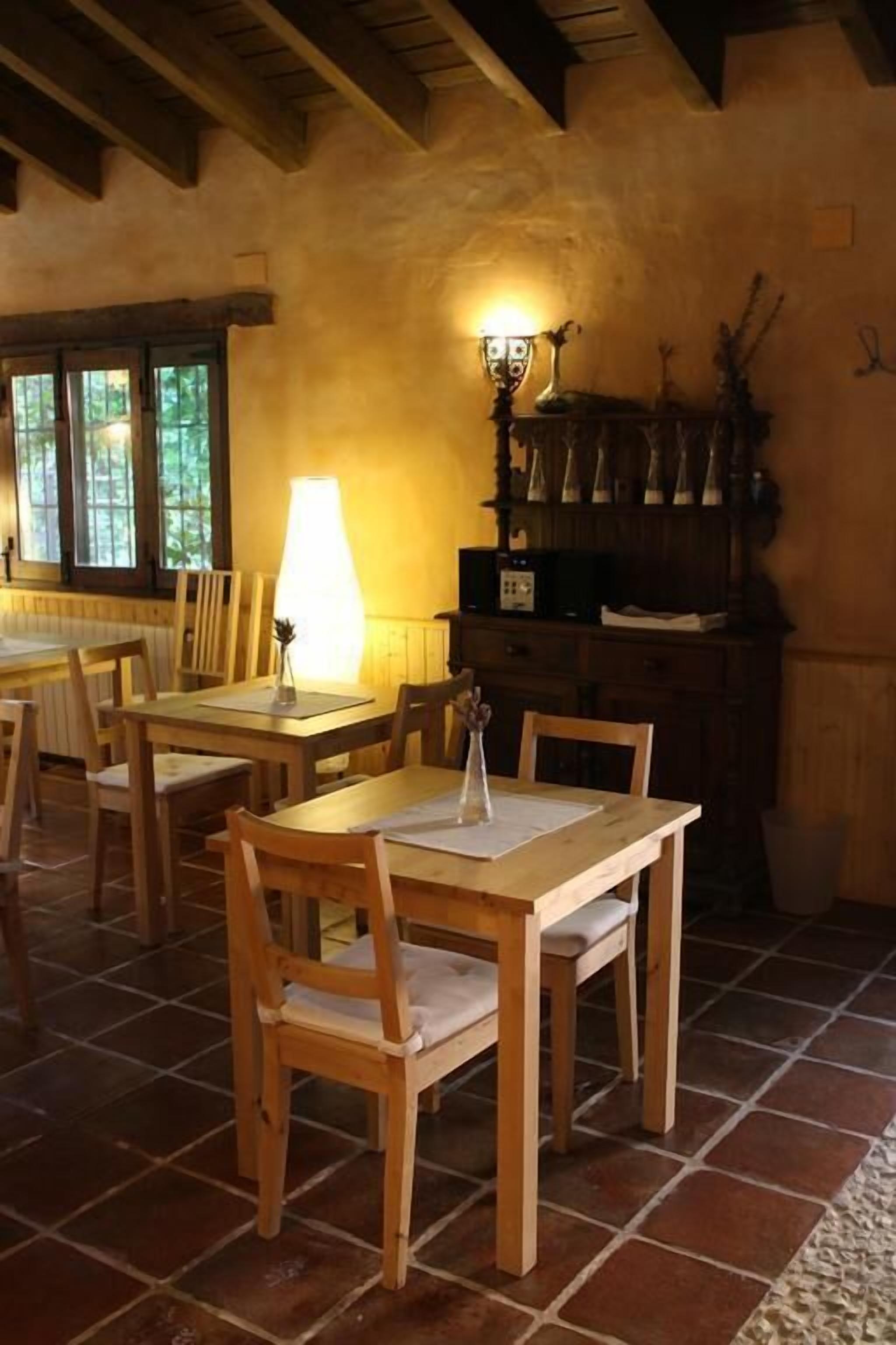 A dining area with wooden tables and chairs, a lamp, and a rustic wall with a shelf.