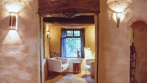 Un intérieur rustique avec un sol en pierre, des poutres en bois et une fenêtre à barreaux. On y trouve deux fauteuils blancs et une petite table sur laquelle est posé un vase.