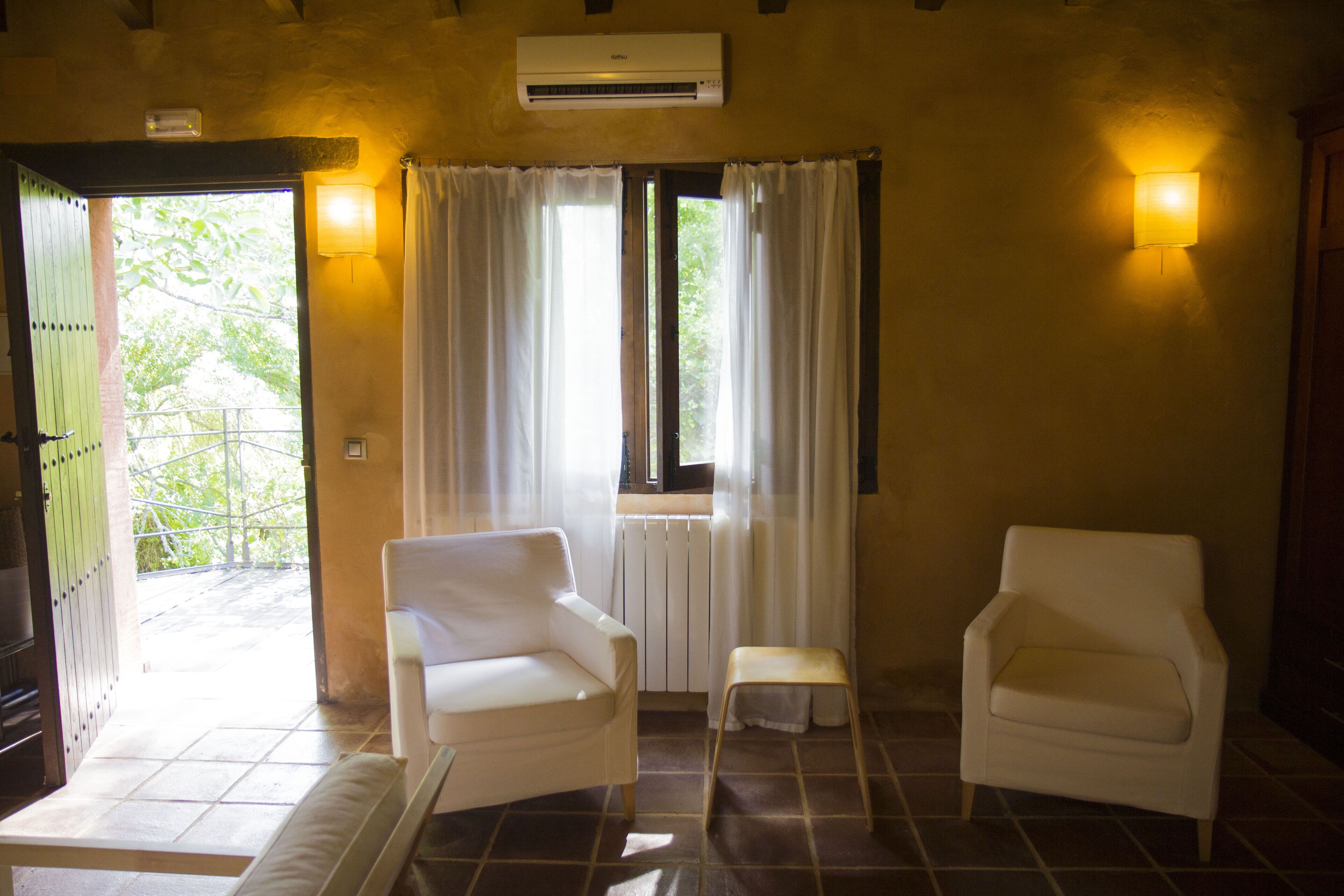 A room with two white chairs, a small stool, a window with curtains, and a wall-mounted air conditioner.