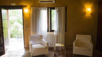 A room with two white chairs, a small stool, a window with curtains, and a wall-mounted air conditioner.