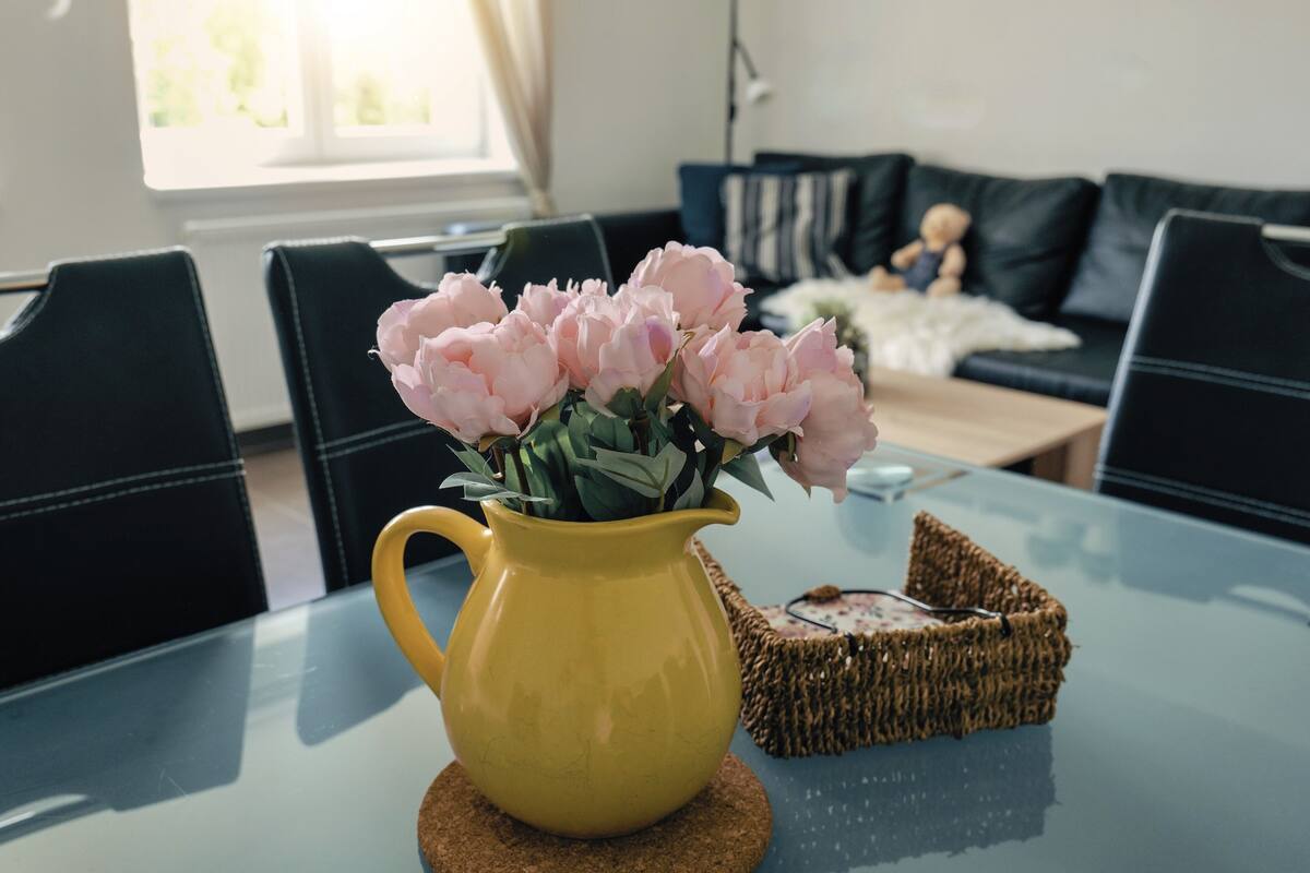 A yellow pitcher with pink flowers on a glass table, surrounded by black chairs and a dark sofa with striped pillows.