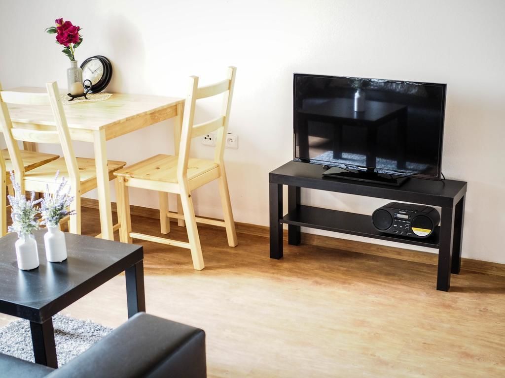 A room with a wooden dining table, two chairs, a black TV stand with a flat-screen TV, and a small black table with two white vases.