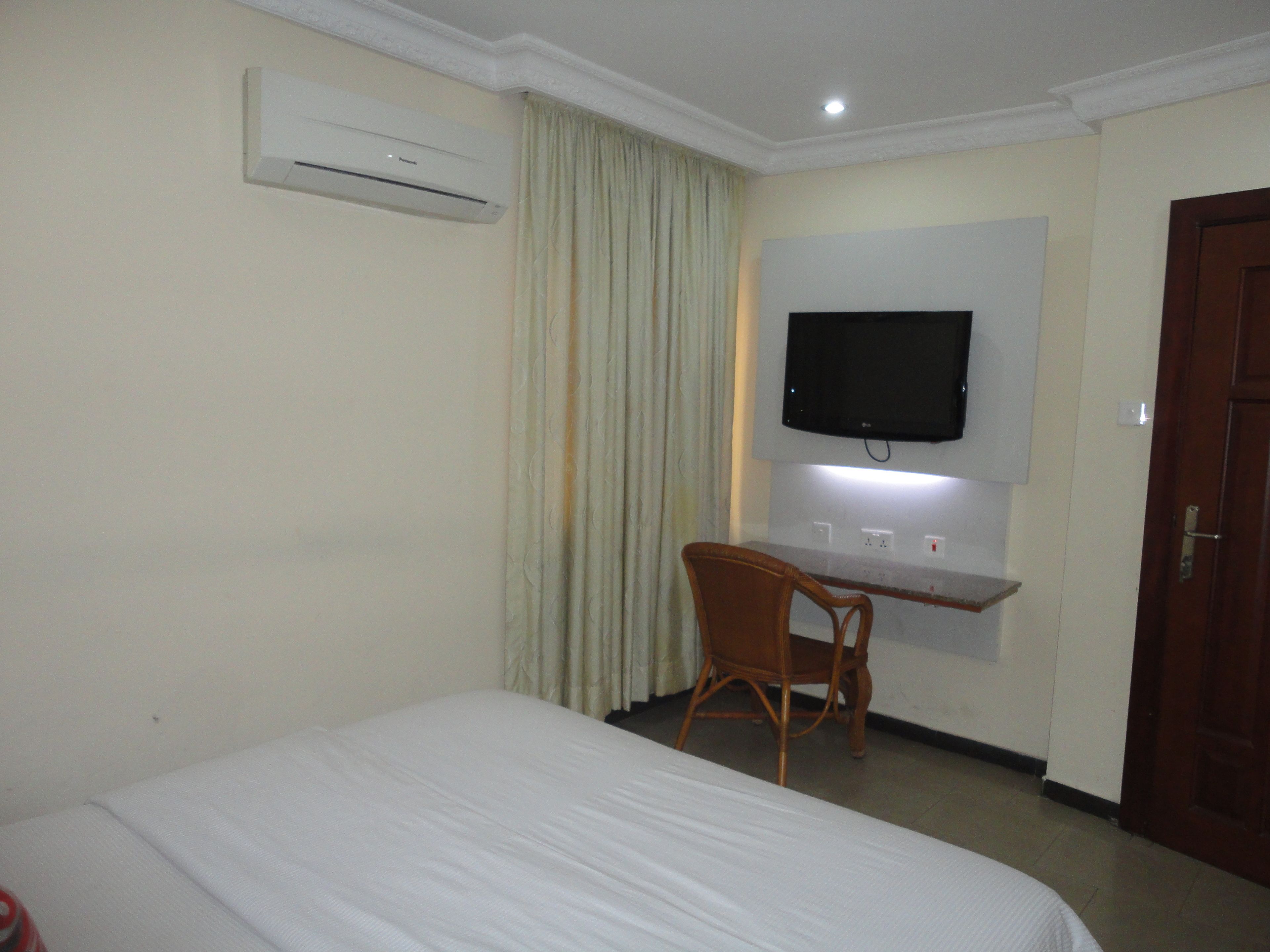 A hotel room with a bed, a desk with a chair, a television mounted on the wall, and an air conditioning unit.