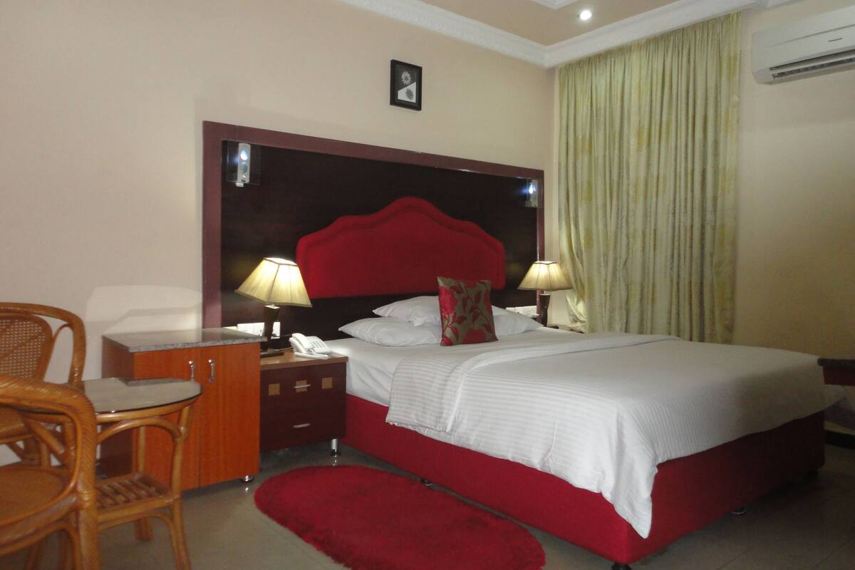 A hotel room with a large bed, two bedside lamps, a nightstand, a telephone, a chair, and a red rug.