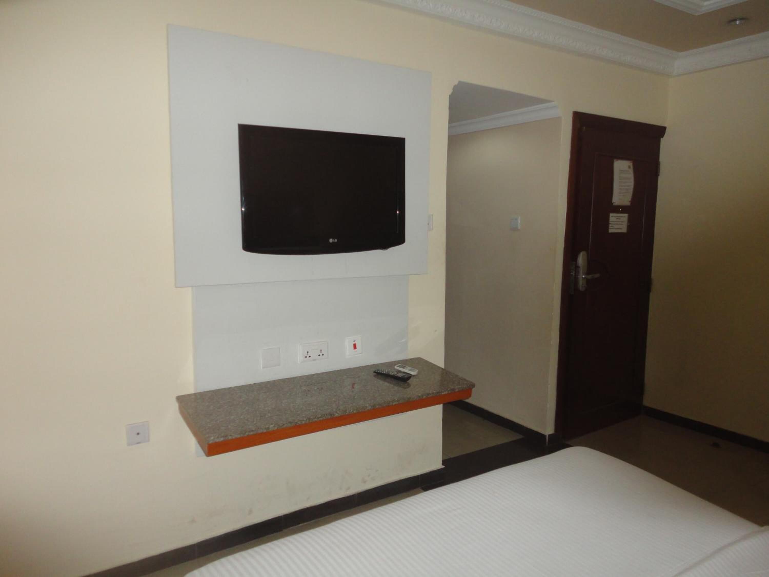 A hotel room with a flat-screen TV mounted on the wall, a small wooden shelf below it, a bed with white bedding, and a door to the right.