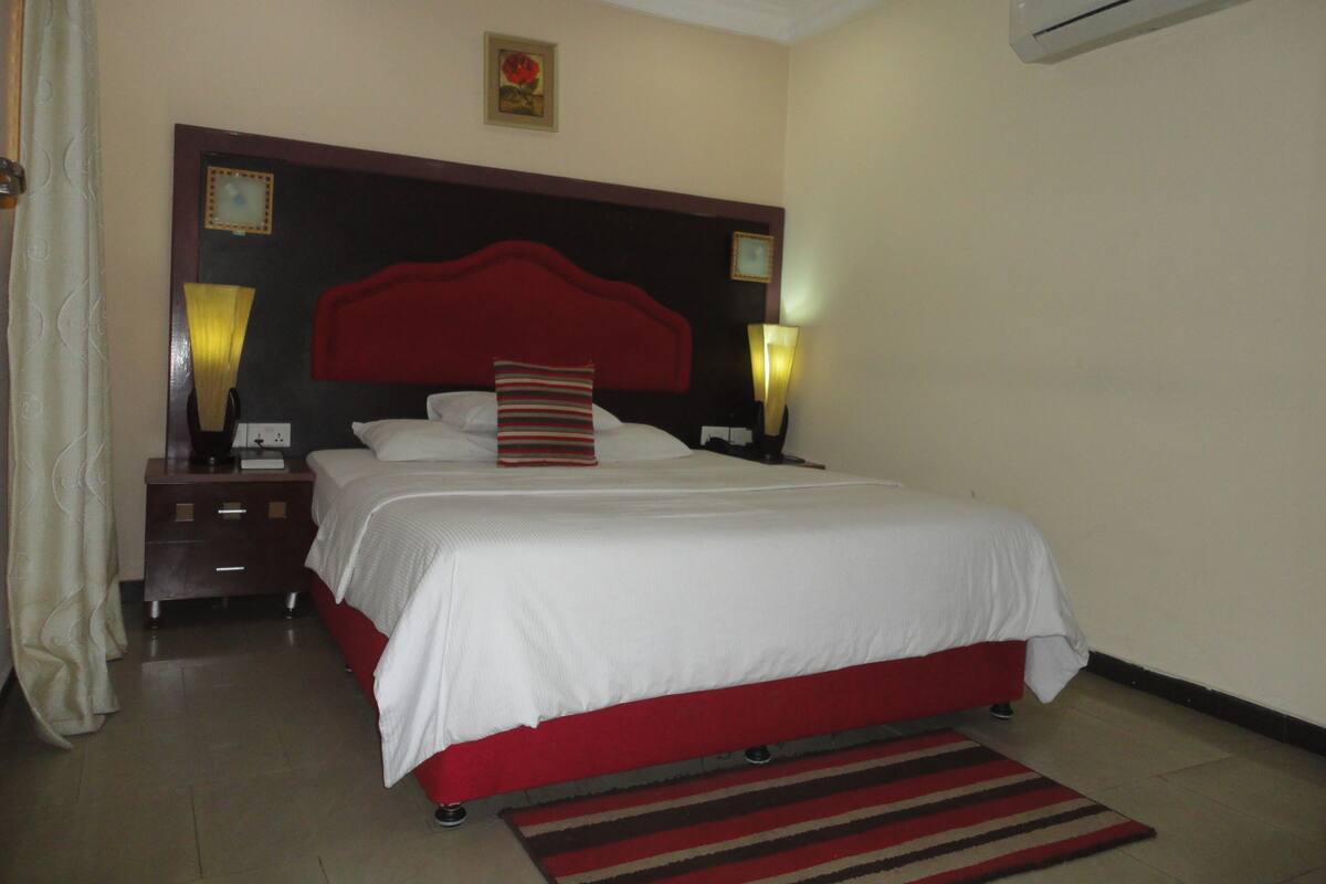 A hotel room with a large bed, a red headboard, two bedside lamps, a nightstand, and an air conditioner on the wall.