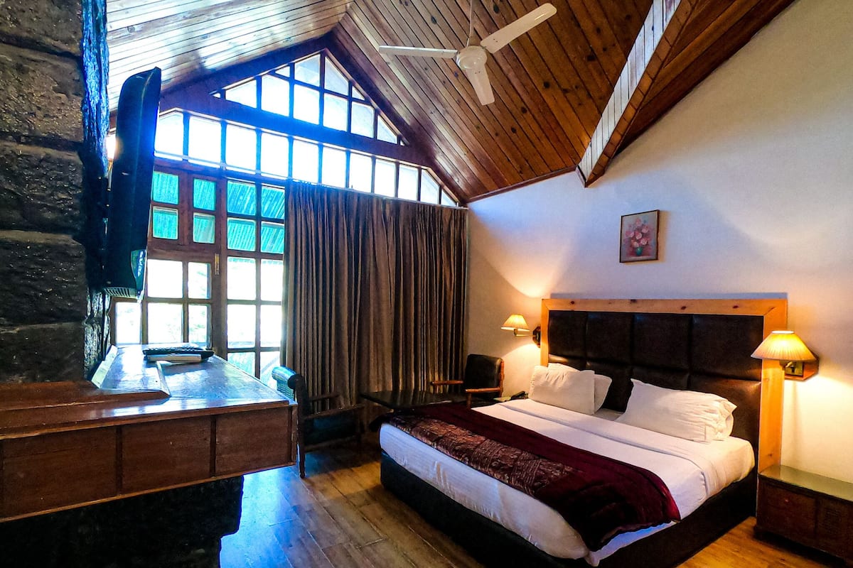 A hotel room with a wooden ceiling, a large bed with white linens, a desk with a chair, and a window with curtains.