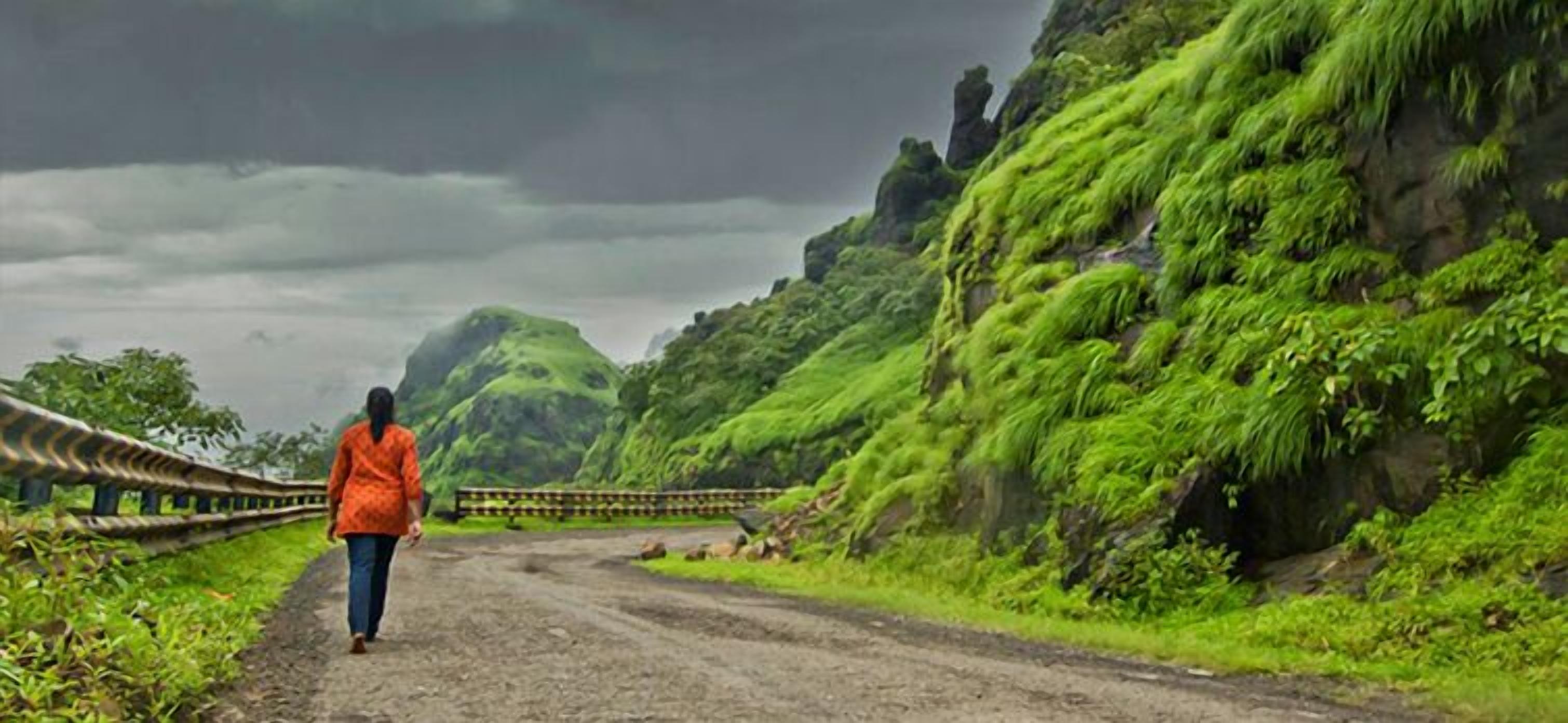 A person walking on a dirt road surrounded by lush green hills and a cloudy sky.