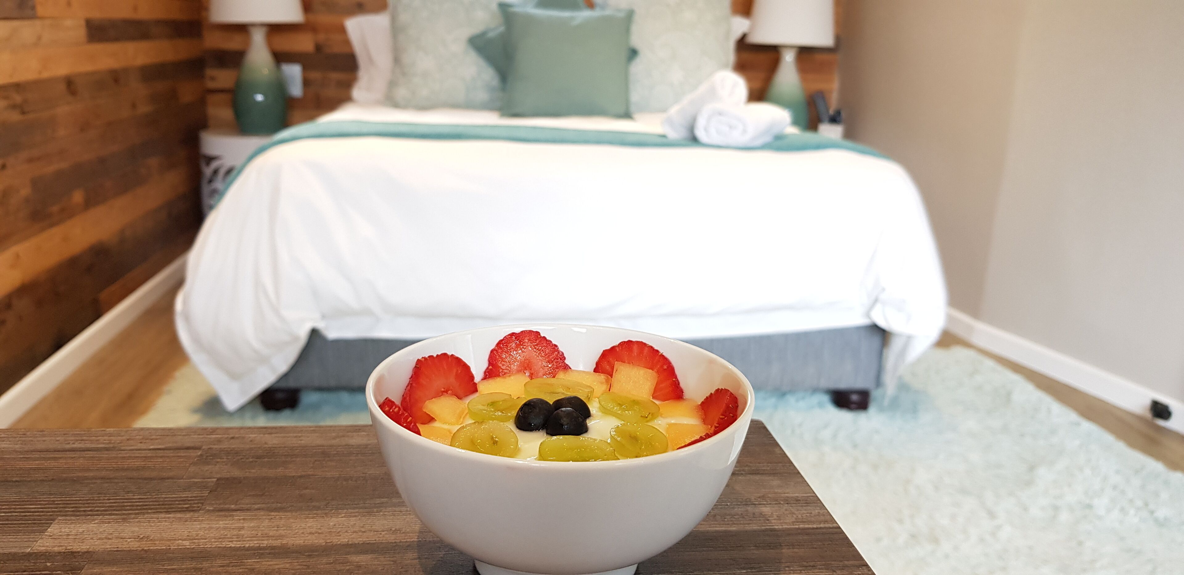 A hotel room with a bed, a bedside table with a bowl of fruit, and a wooden wall.