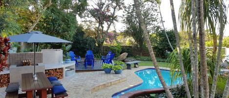 A backyard with a pool, blue chairs, a barbecue area, and a wooden deck.