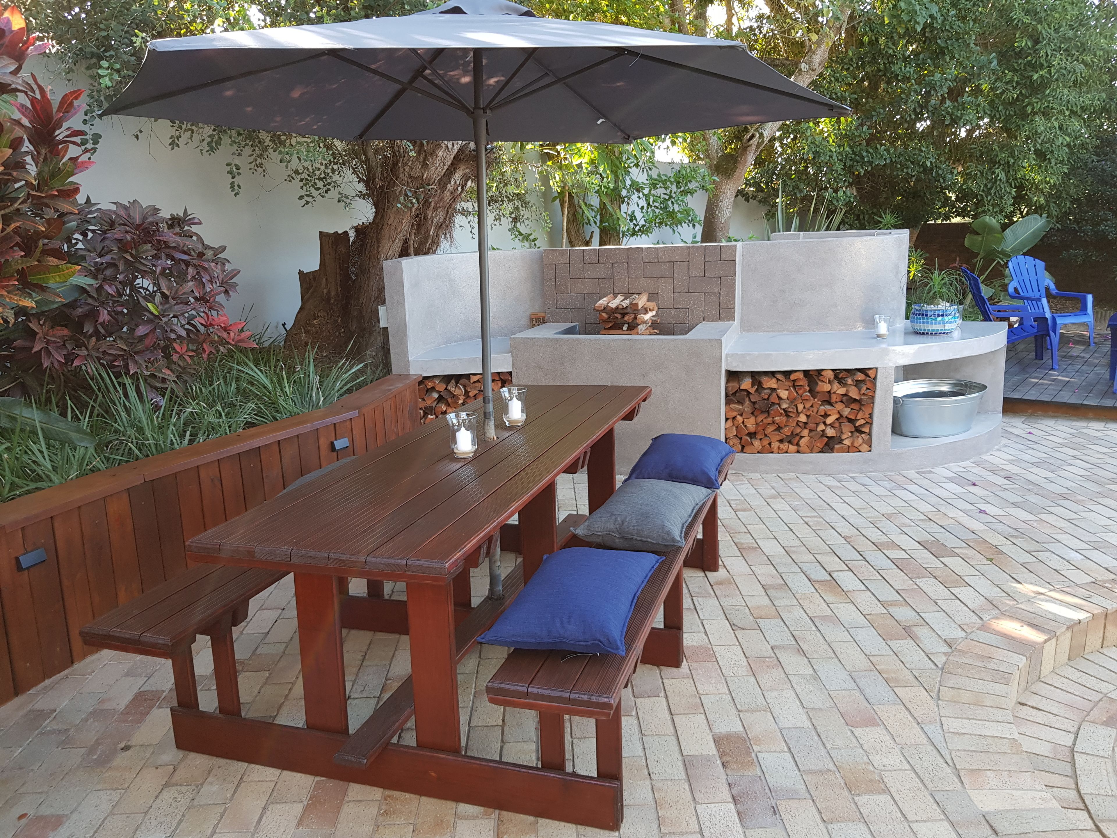 An outdoor patio with a wooden table and benches, a large umbrella, and a stone fireplace.