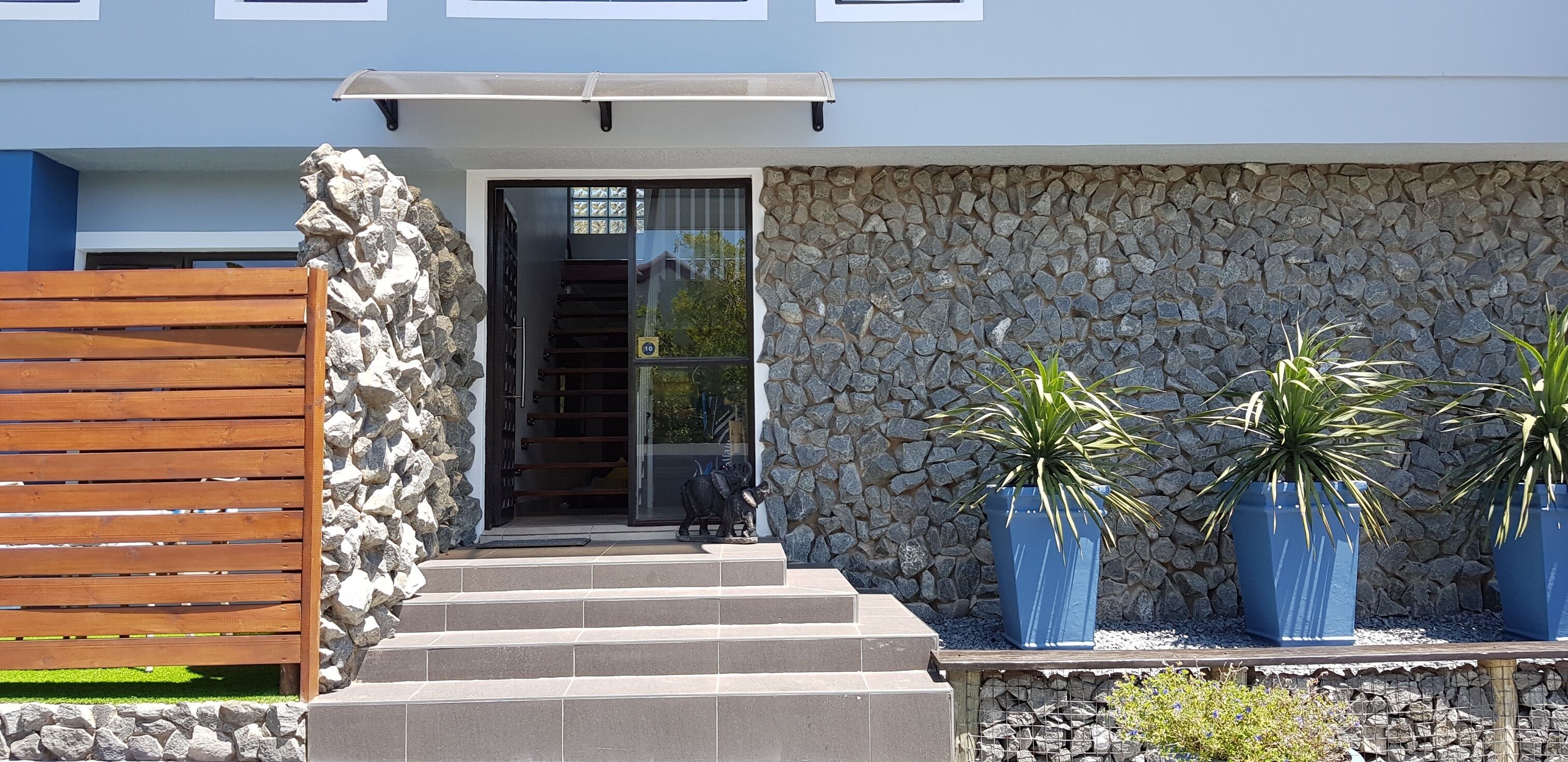 A modern building entrance with a stone wall, a wooden fence, and three large potted plants.